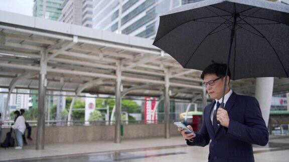 自信的亚洲商人办公室职员在雨中拿着黑色雨伞使用智能手机帅哥商务人士工作在城市办公区在恶劣的天气