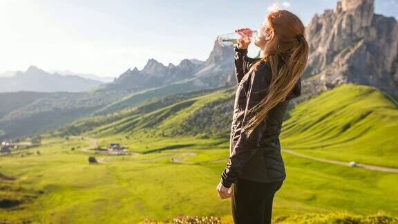 一名女子站在山顶俯瞰山谷用可重复使用的水瓶喝水