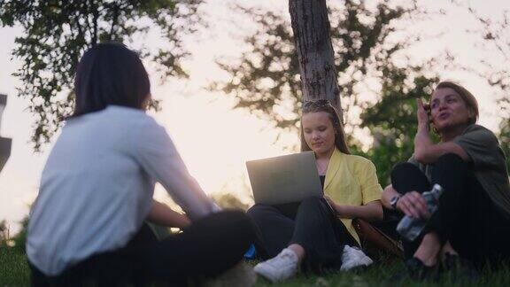 三个多民族的女商人坐在公园的草地上聊天其中一个在笔记本电脑上工作