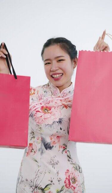 中国亚洲妇女在中国新年期间双手拿着红色和粉红色的购物袋白色背景穿着旗袍白色花卉图案连衣裙