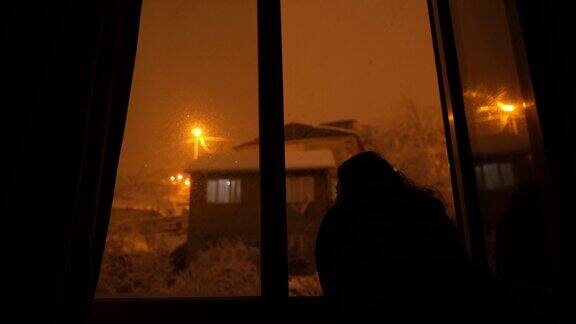 女性剪影从窗口观看雪花飘落的夜晚