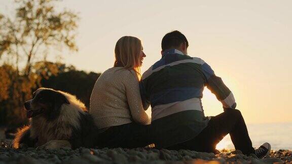 日落时分一对年轻的多民族夫妇和一只狗坐在海滩上一个人把小石子扔进水里