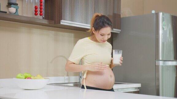 亚洲美女怀孕站在家里喝牛奶迷人的孕妇站在子宫里抚摸宝宝喝鲜奶健康饮料在厨房里感受幸福