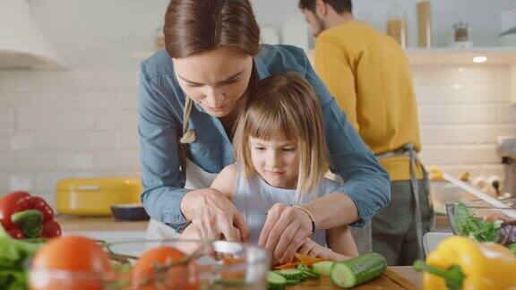 在厨房:妈妈可爱的小女儿一起做健康晚餐妈妈教小女孩健康的习惯和如何切蔬菜做沙拉可爱的孩子帮助她美丽的父母