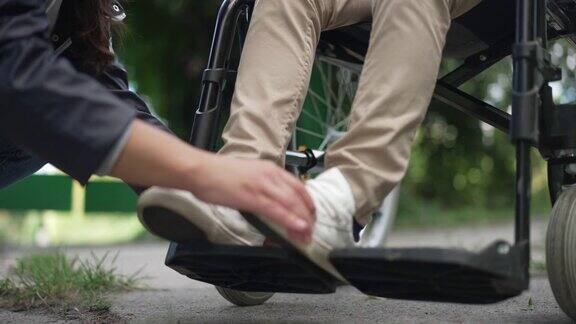 一位无法辨认的妇女正在调整轮椅上残疾儿童的双腿年轻的白人母亲在夏日公园的户外照顾瘫痪的儿子