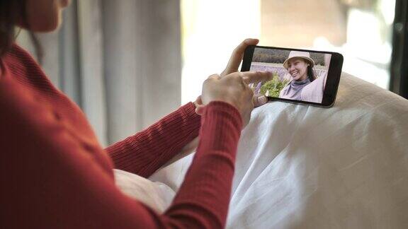 年轻女子拿着智能手机摄像头与躺在家里床上的朋友视频聊天