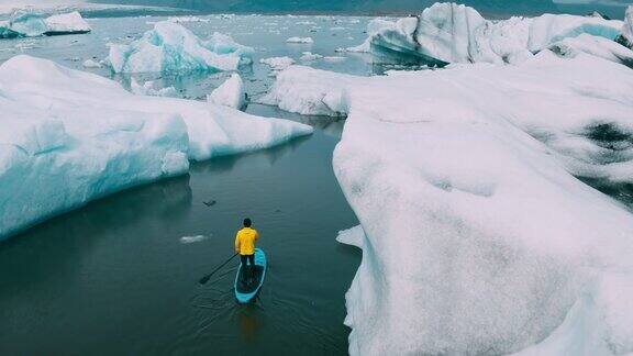 年轻的冒险男子划着桨立桨在冰岛冰川湖的冰山上冲浪