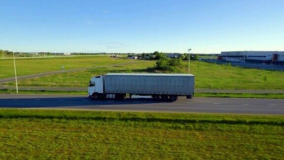 白色半挂车与货运拖车在高速公路上移动的鸟瞰图背景是仓库和农村美丽的晴天