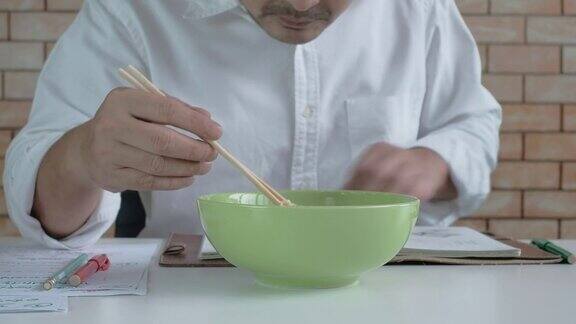 一名亚洲男子一边看书一边吃着绿色碗里的方便面