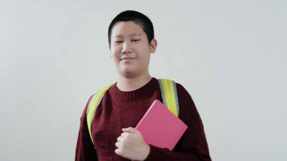 快乐的亚洲男生穿着红色毛衣拿着粉红色的书看着灰色背景的相机生活方式