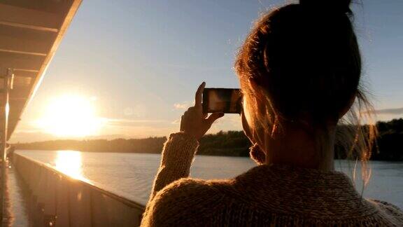 一名女子在游轮甲板上用智能手机拍摄日落剪影
