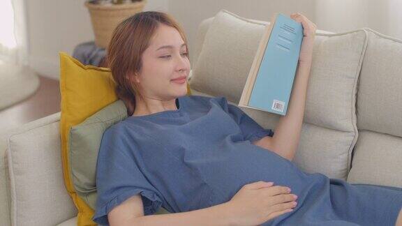 快乐的亚洲孕妇在家里看书轻轻抚摸她的肚子怀孕分娩