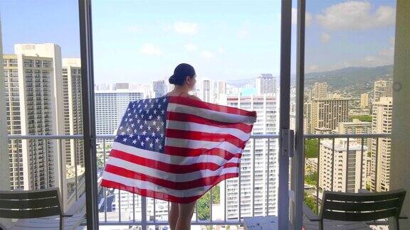 一个拿着美国国旗的女人站在阳台上看着夏威夷檀香山的怀基基海滩4k慢镜头60帧秒