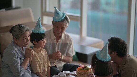 亚洲华人多代家庭闭上眼睛在生日蛋糕前许下生日愿望然后在客厅吹生日蜡烛庆祝儿子生日