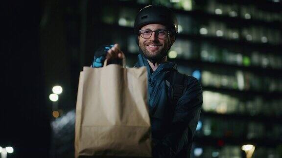 一个年轻的快递员在一个晚上的城市中心在镜头前对他的工作满意地微笑着
