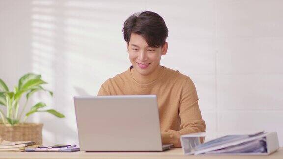 企业家英俊的商业亚洲人穿休闲衬衫在书桌上工作桌上微笑和幸福与笔记本电脑在家里有吸引力的自由职业者在家庭办公室在线销售营销