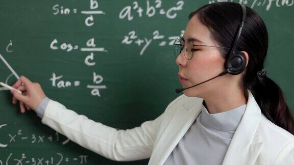 年轻的亚洲女教师手持粉笔戴着耳机与学生看着摄像机进行视频会议女教师在课堂上培训数学黑板在线课程