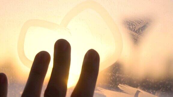 女孩女人用手在日落时在冻结的窗户上画了一颗心