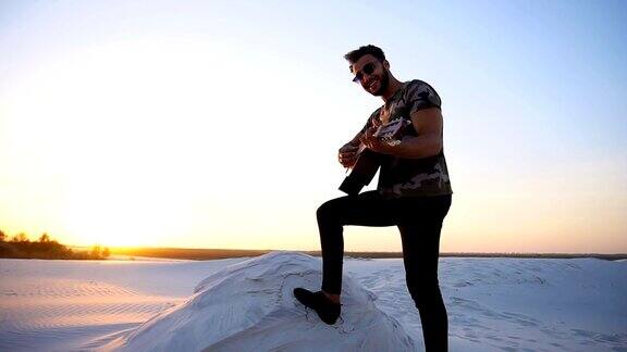 在一个温暖的夜晚一个英俊的阿拉伯人正在弹吉他站在沙漠中的小山丘上夕阳西下