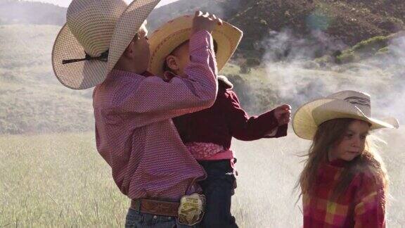 一个年轻的牛仔把一顶帽子放在一个小牛仔身上而一个年轻的女孩在看篝火