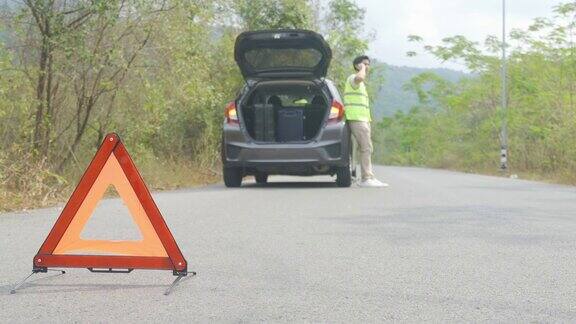 故障三角标志在道路上与担心的亚洲人通过手机与保险或汽车服务中心公司后汽车故障在乡村道路上的背景
