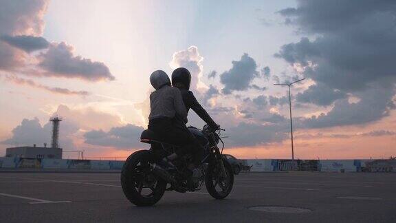 年轻夫妇戴着头盔骑着摩托车在城市里以美丽的日落天空为背景慢镜头