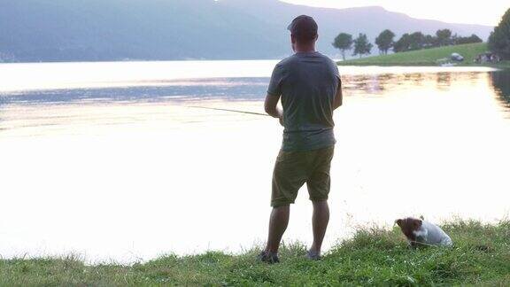 夏天日落时渔夫在平静的水面上旋转滑梯年轻人站在一个平静的大湖岸边拿着钓竿钓鱼镜头反射狗杰克罗素梗