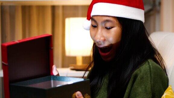 快乐的亚洲女孩穿着绿色毛衣和圣诞老人帽坐在沙发上打开圣诞礼盒在家里节日