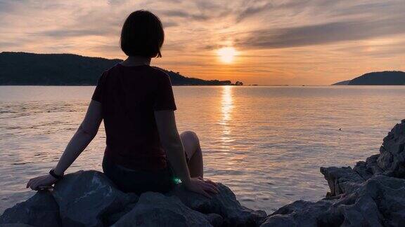 这名年轻女子坐在五彩缤纷的日落背景下自拍用智能手机拍照然后发送给朋友美丽的海上景观