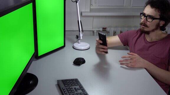 一个年轻人正坐在办公室的双绿屏幕电脑前使用手机