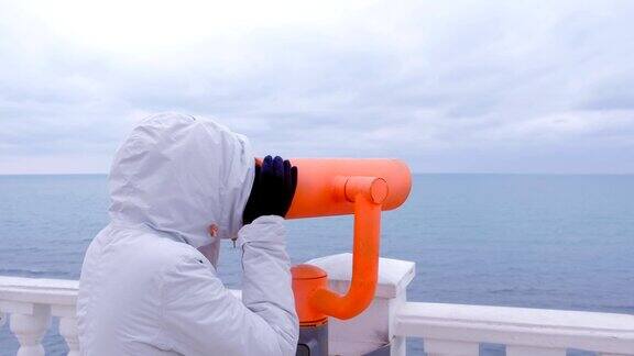 女人在白色夹克和兜帽在双筒望远镜看海滨海景侧视图