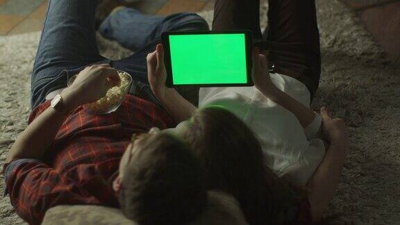 年轻夫妇晚上躺在家里的地板上看绿色屏幕的平板电脑休闲的生活方式