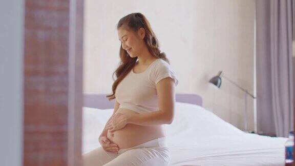 快乐怀孕的亚洲女人坐在床上抱着和抚摸着自己的大肚子年轻女人的怀孕享受和未来的生活放松在家里母亲和怀孕柔软的焦点