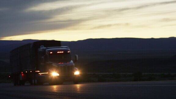 一辆带有前灯的半挂车在黄昏时驾驶在科罗拉多西部的高沙漠背景是科罗拉多国家纪念碑的剪影