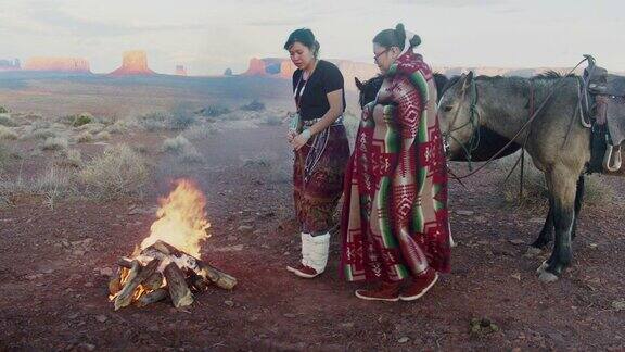 印第安姐妹一个在打鼓裹着传统的纳瓦霍毯子在篝火旁取暖他们身后的马标志性的纪念碑日落