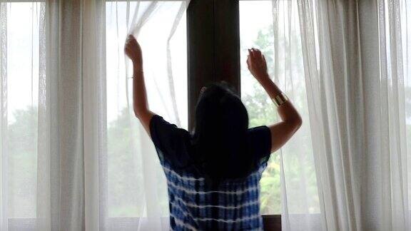 亚洲妇女在早晨打开窗帘和窗户看外面的阳台度假暑假假期