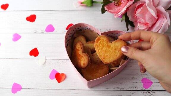 情人节的心形饼干在蓝色盒子里粉红色的玫瑰和心形放在白色的木桌上女性的手把饼干放到盒子里