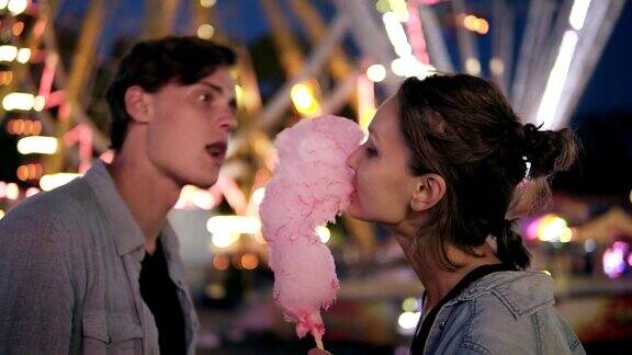 一对可爱的年轻夫妇在游乐场花时间在一起吃粉色的棉花糖爱的夫妻约会模糊的游乐场背景