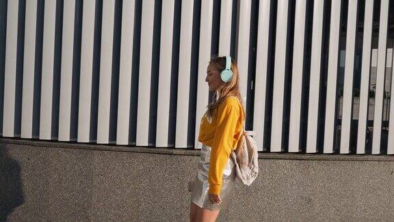 年轻可爱迷人的年轻女孩的肖像在城市背景听音乐耳机穿着黄色衬衫和银色裙子的女人