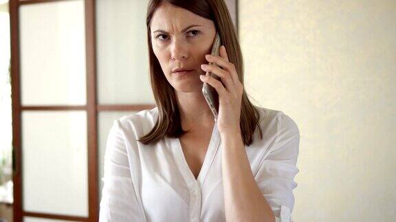 不经意的年轻女子在白衬衫站在客厅打电话收到坏消息心烦意乱