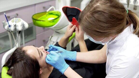 年轻的女病人拿着镜子在看牙医检查手术的结果年轻女牙医戴着手套和面罩带着工具走近