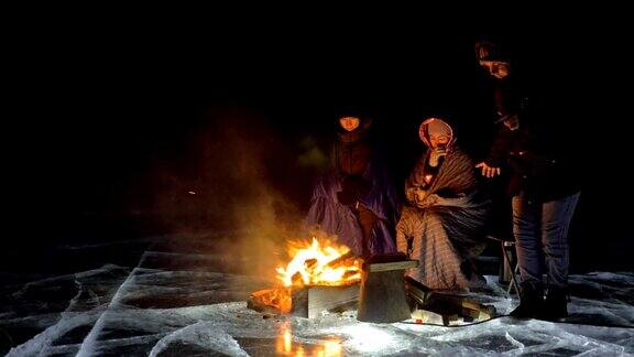 三个旅行者在夜间的冰面上着火营地在冰上帐篷紧挨着火贝加尔湖附近有一辆车人们围着篝火取暖穿着睡袋
