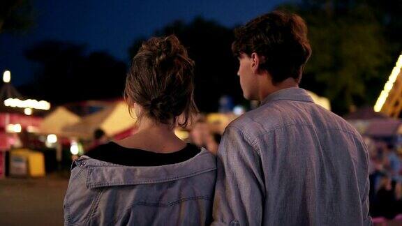 一个年轻的约会夫妇在晚上漫步在游乐场吃棉花糖的背影两人都穿着相似的蓝色衬衫游乐园情侣拥抱
