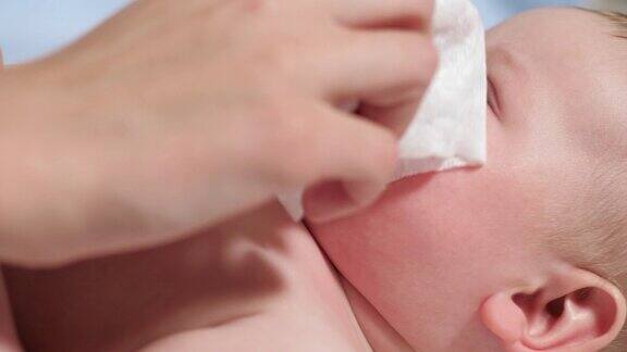 妈妈用湿纸巾擦拭宝宝的鼻子孩子躺在蓝色背景的白毯子上妈妈用湿布擦他的鼻子慢动作