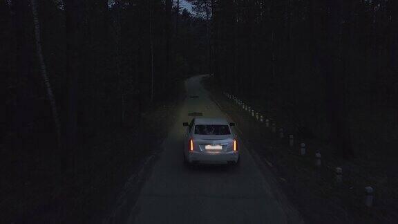 空中跟随视图白色豪华轿车凯迪拉克Cts驾驶在黑暗的乡村道路在夏天的森林在晚上从无人驾驶汽车氙气前灯行驶在夜晚的道路通过黑暗的森林