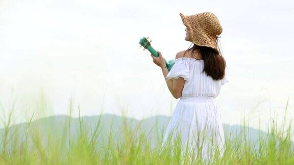 亚洲快乐的女人在白色的衣服弹奏尤克里里吉他在绿色的草地与山的背景在夏天音乐家美女放松与乐器乡村的生活方式