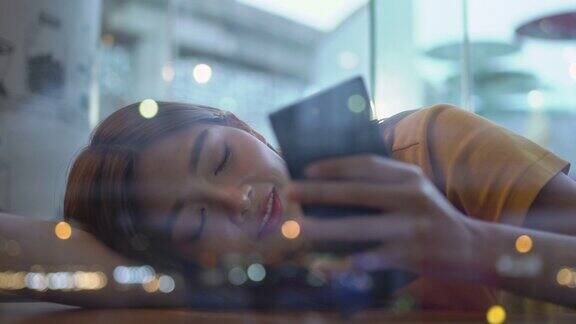 4K孤独年轻美丽的亚洲女人坐在窗前躺在桌子上晚上在咖啡馆用智能手机上网美丽的少女望着窗外灯火通明的城市街道