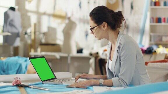 美丽的女性时装设计师坐在她的办公桌上用绿色屏幕的笔记本电脑工作她的工作室是阳光色彩丰富的面料和人体模型在那里
