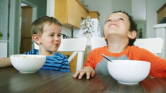 两个白人小男孩(三岁和五岁)坐在餐桌旁一边吃着碗通心粉和奶酪意大利面一边笑着做着傻乎乎的动作
