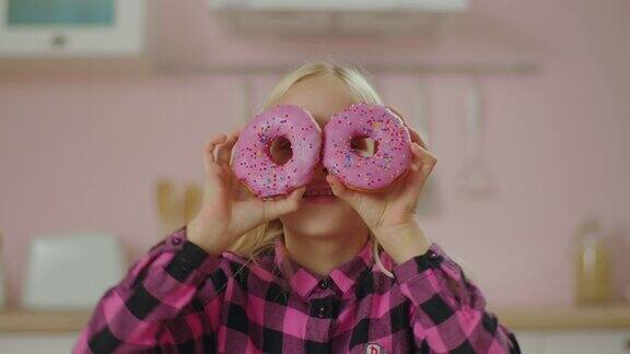 可爱的金发小女孩穿着粉红色的衬衫手里拿着两个粉色的甜甜圈粉红色的甜甜圈的眼睛糖果的有趣概念
